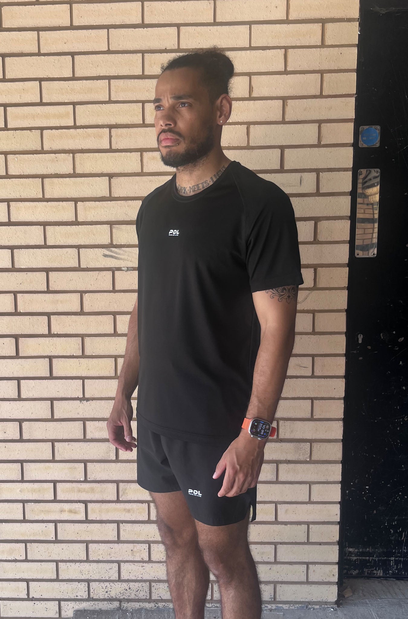 Man wearing a black t-shirt and shorts standing against a brick wall.
